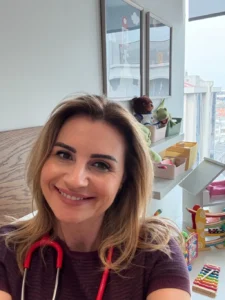 Uzm. Dr. İrem Oral’ın samimi gülümsemesi ve stetoskopu ile, arka planda çocuk oyun köşesinin yer aldığı kliniğinde hasta karşılamaya hazır olduğu an.